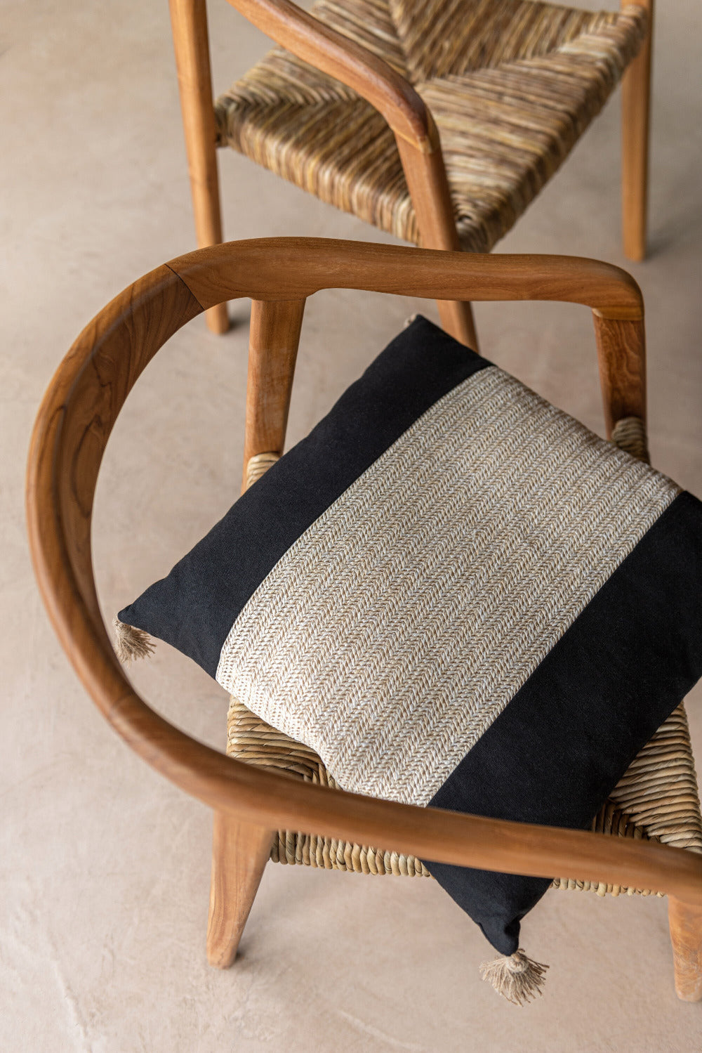 Zwart sierkussen met centrale beige geweven strook en franjes, geplaatst op een houten stoel met gevlochten zitting, met een tweede soortgelijke stoel op de achtergrond op een beige vloer