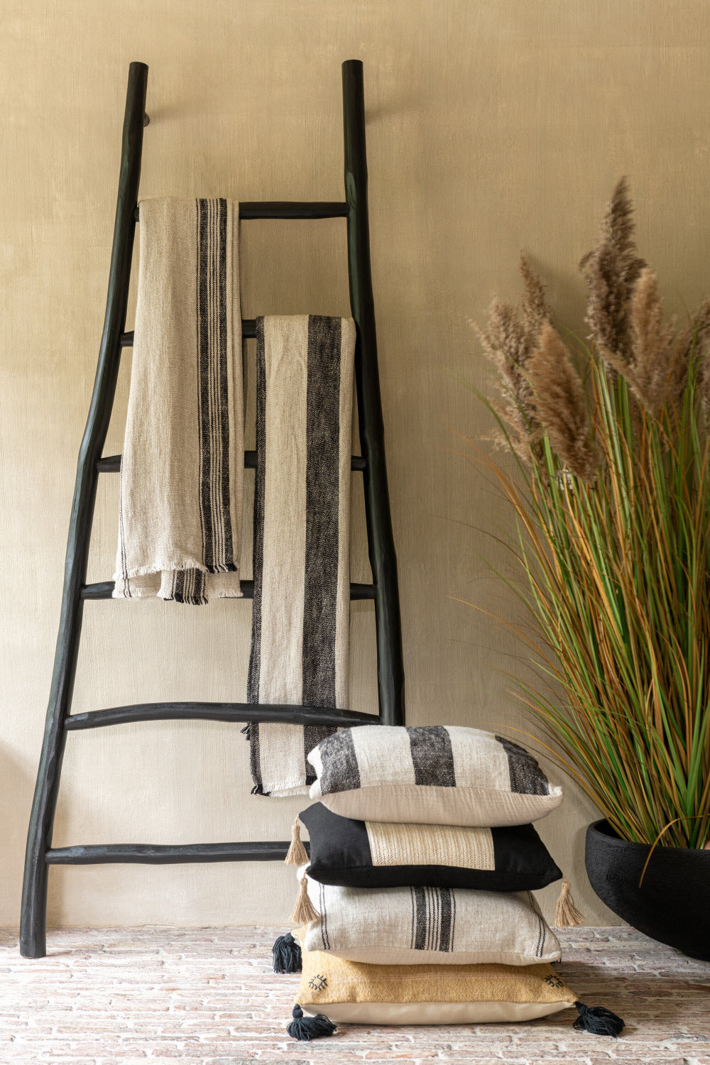 Gestileerd hoekje met zwarte houten ladder tegen een beige muur, twee beige plaids met zwarte strepen over de sporten, daarnaast een stapel van vier sierkussens in natuurlijke tinten met franjes, en een zwarte pot met siergras