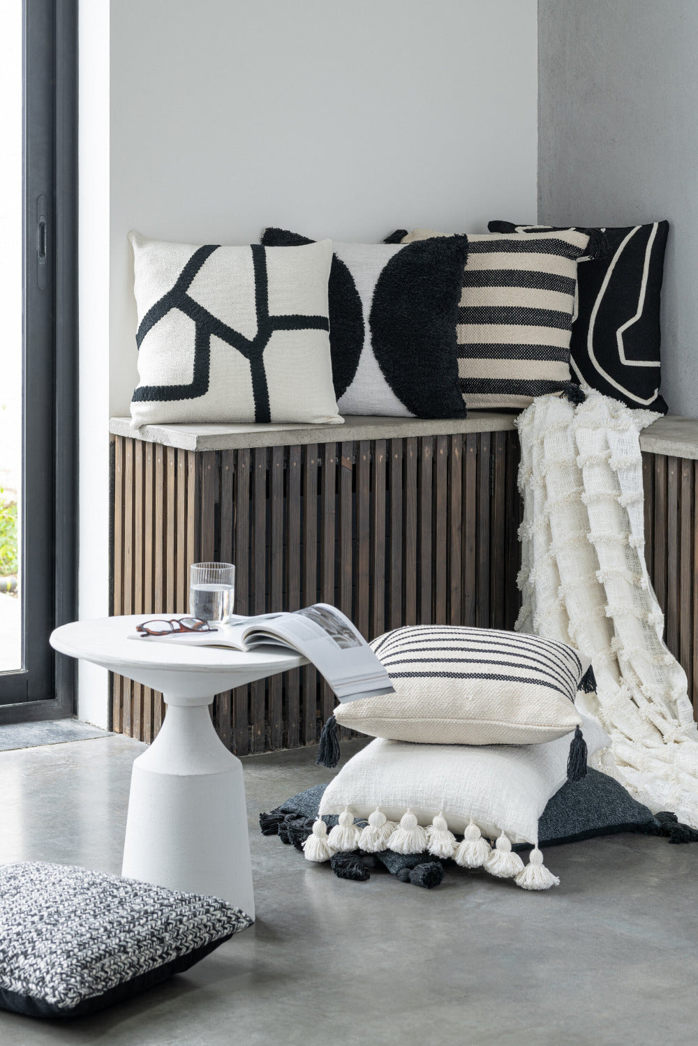 Scandinavisch interieur met zwart-witte sierkussens, witte plaid met franjes en minimalistische ronde bijzettafel met boek en bril