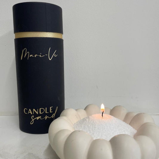 Mari-Ve koker met Candle Sand naast een brandend lontje in wit decoratief schaaltje – sfeervolle presentatie van kaarsenzand in gebruik