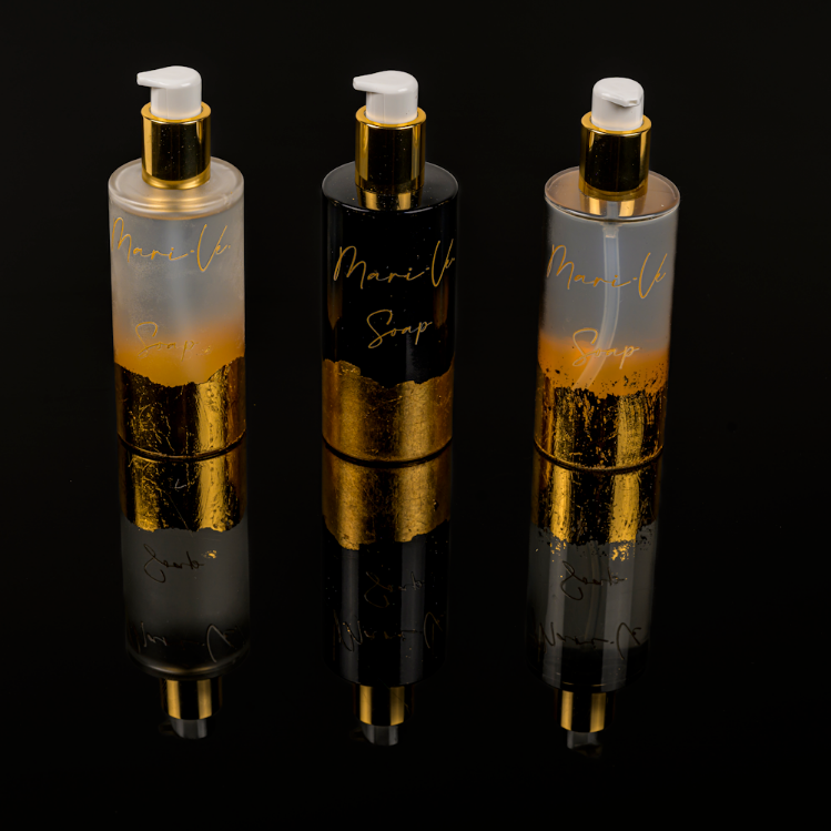 Drie stijlvolle zeepdispensers uit de Mari-Ve collectie, met transparante, zwarte en gemarmerde flacons voorzien van luxe gouden details en reflectie op een glanzend zwart oppervlak.